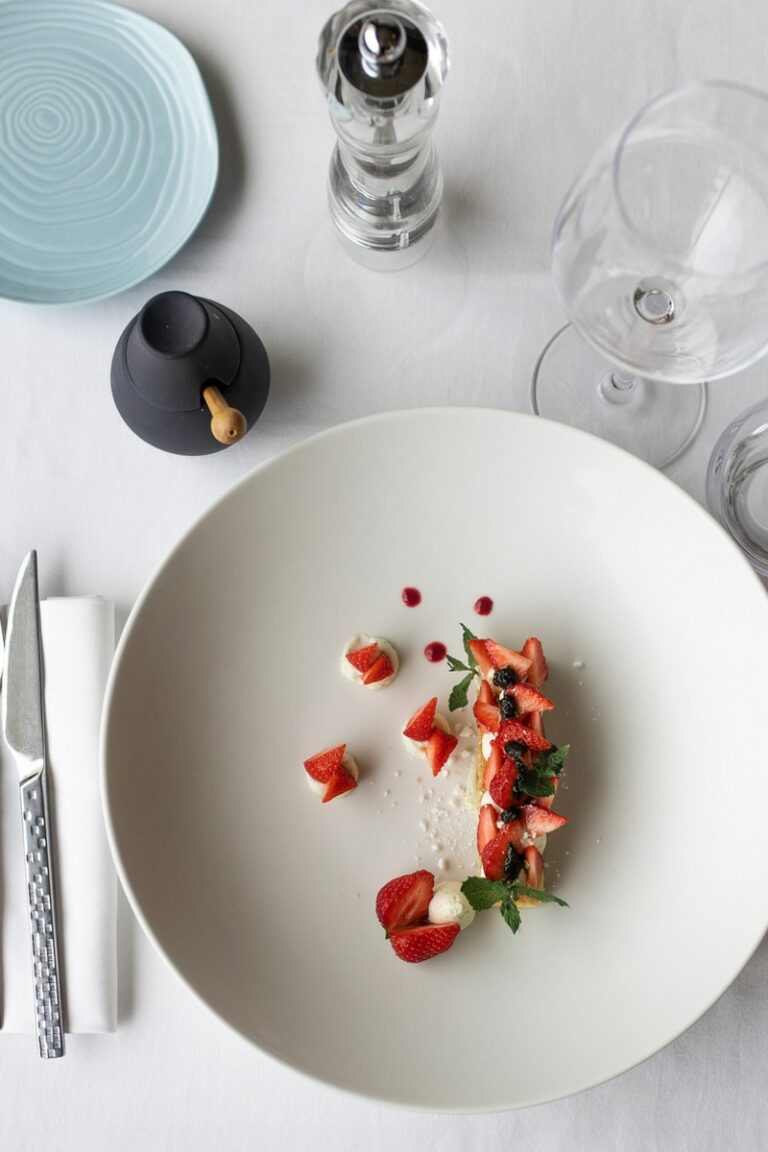 Photographie artistique de dessert, où des fraises fraîches sont harmonieusement disposées avec de la crème et des touches de sauce rouge sur une assiette blanche épurée
