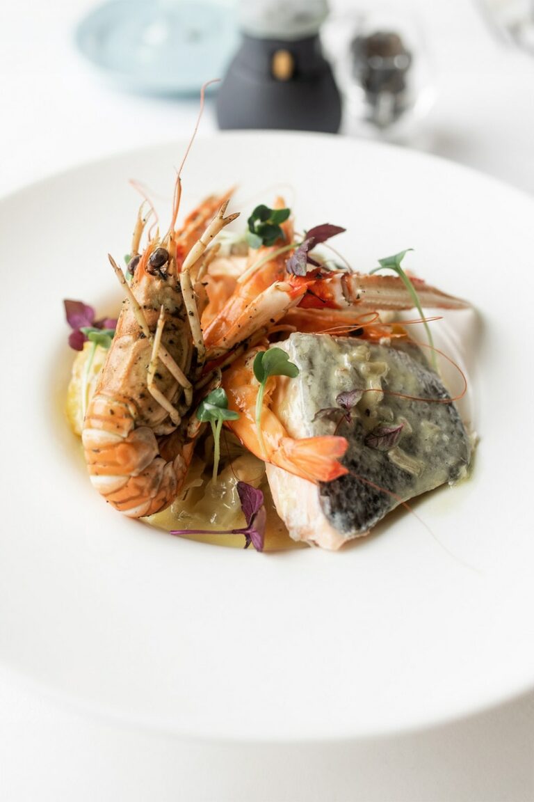 Photographie culinaire d'un plat de fruits de mer élégamment présenté, avec des langoustines joliment disposées au-dessus d'une préparation de poisson et de légumes, capturée dans l'ambiance lumineuse d'un restaurant