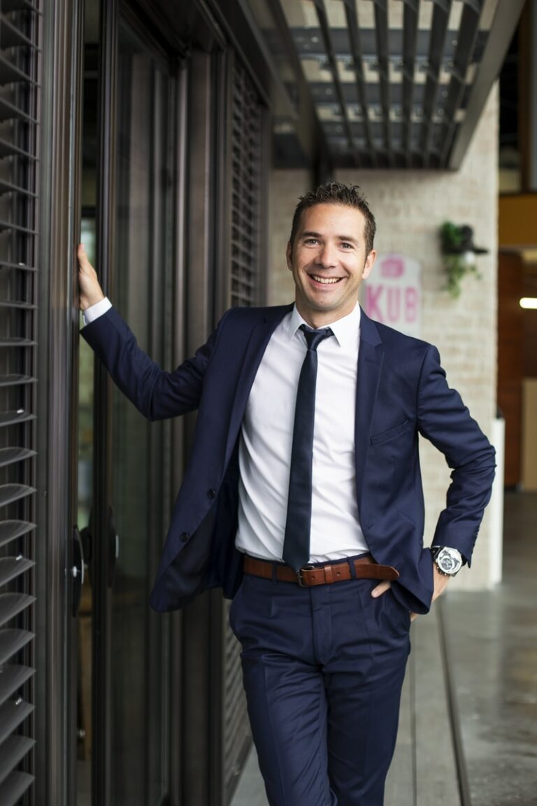 Un jeune entrepreneur souriant en costume bleu marine, chemise blanche et cravate noire, debout à l'entrée d'une véranda