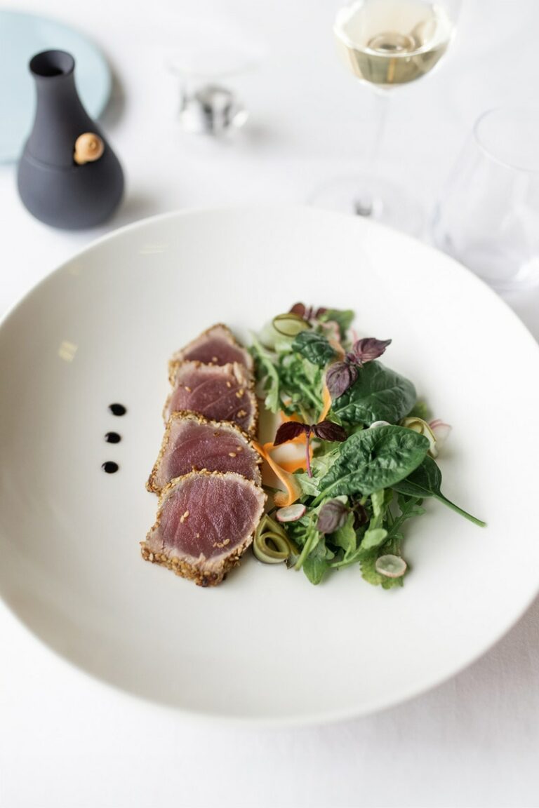 Photographie de cuisine raffinée présentant des tranches de thon mi-cuit en croûte de sésame sur un lit de légumes verts et herbes fraîches, dans une assiette blanche moderne