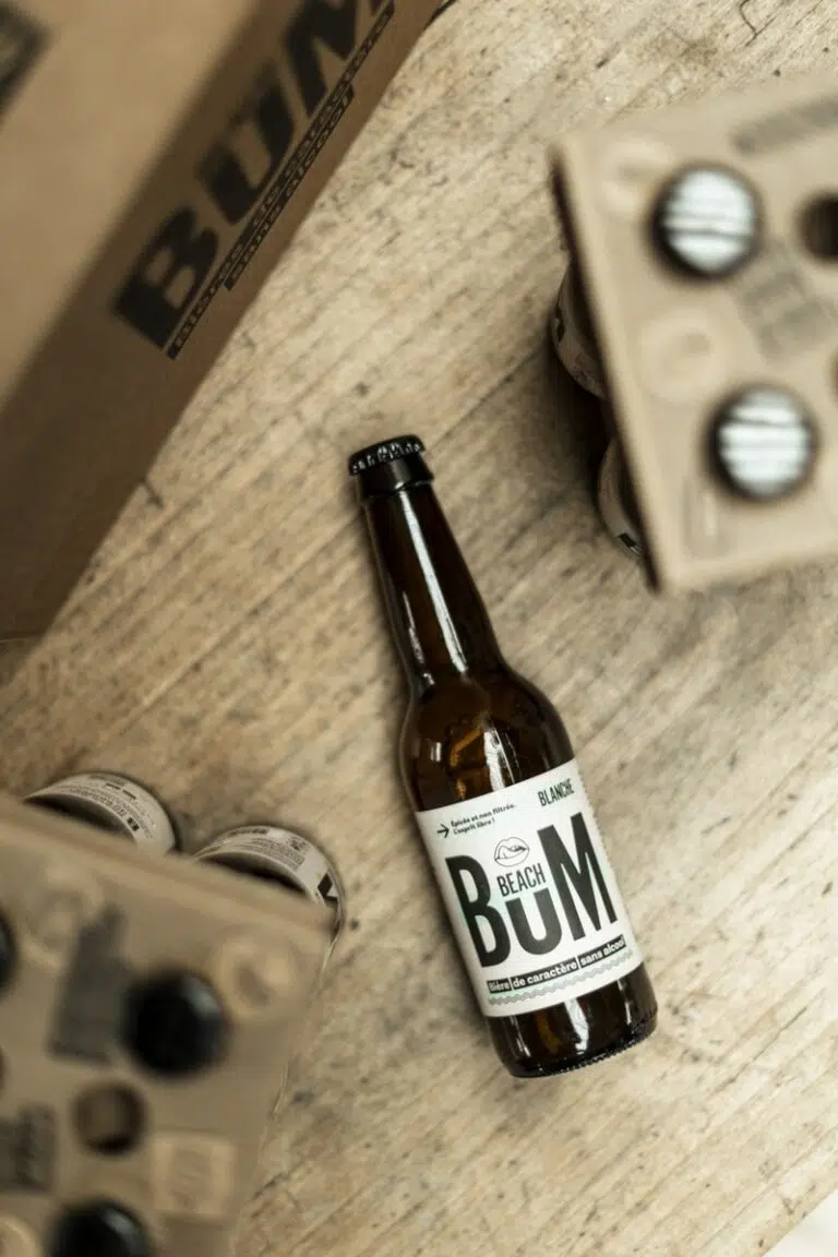 Une image d'une bouteille de bière Beach Bum Blanche posée horizontalement sur une surface en bois, entourée de son emballage en carton.