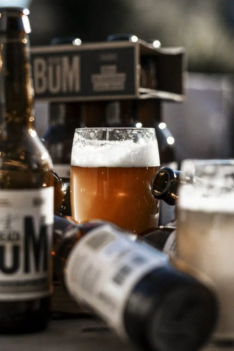 Une image d'un verre de bière de couleur ambre clair avec une écume épaisse et blanche, entouré de plusieurs bouteilles de bière brunes étiquetées 'BUM', faisant partie de la marque Beach Bum. La scène a une ambiance chaude et conviviale avec un fond flou d'un milieu extérieur.
