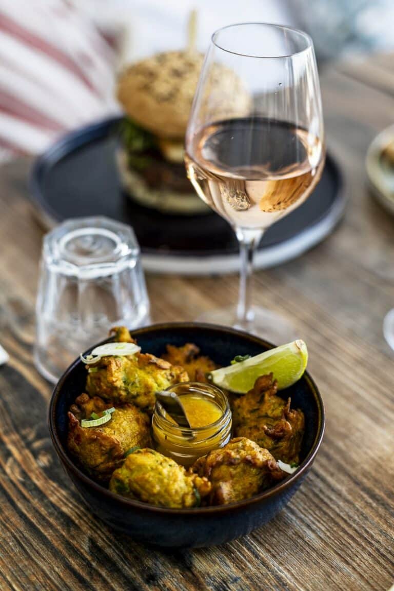 Un bol d’accras de morue avec une tranche de citron vert et une sauce, accompagné d’un verre de vin rosé, sur une table en bois rustique.