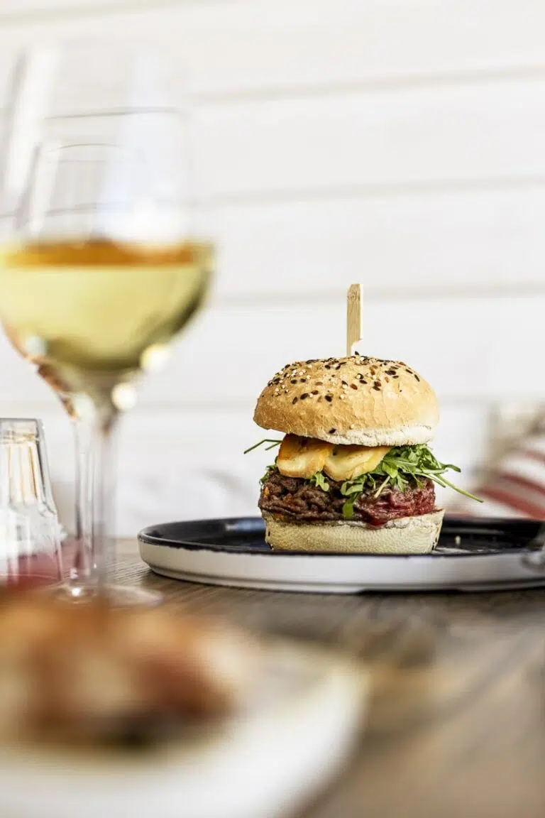 Un burger gourmet garni de steak, fromage grillé, roquette et sauce, servi sur une assiette noire avec un verre de vin blanc en premier plan.