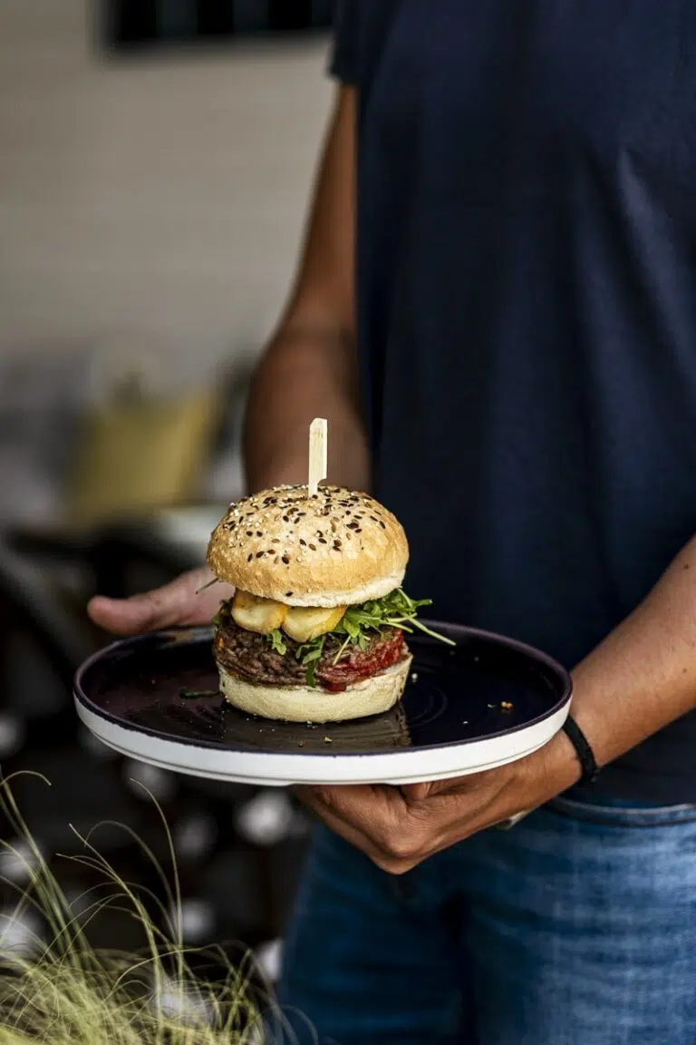Une personne tenant une assiette avec un burger garni de fromage, roquette et steak haché, en extérieur sur une terrasse estivale.