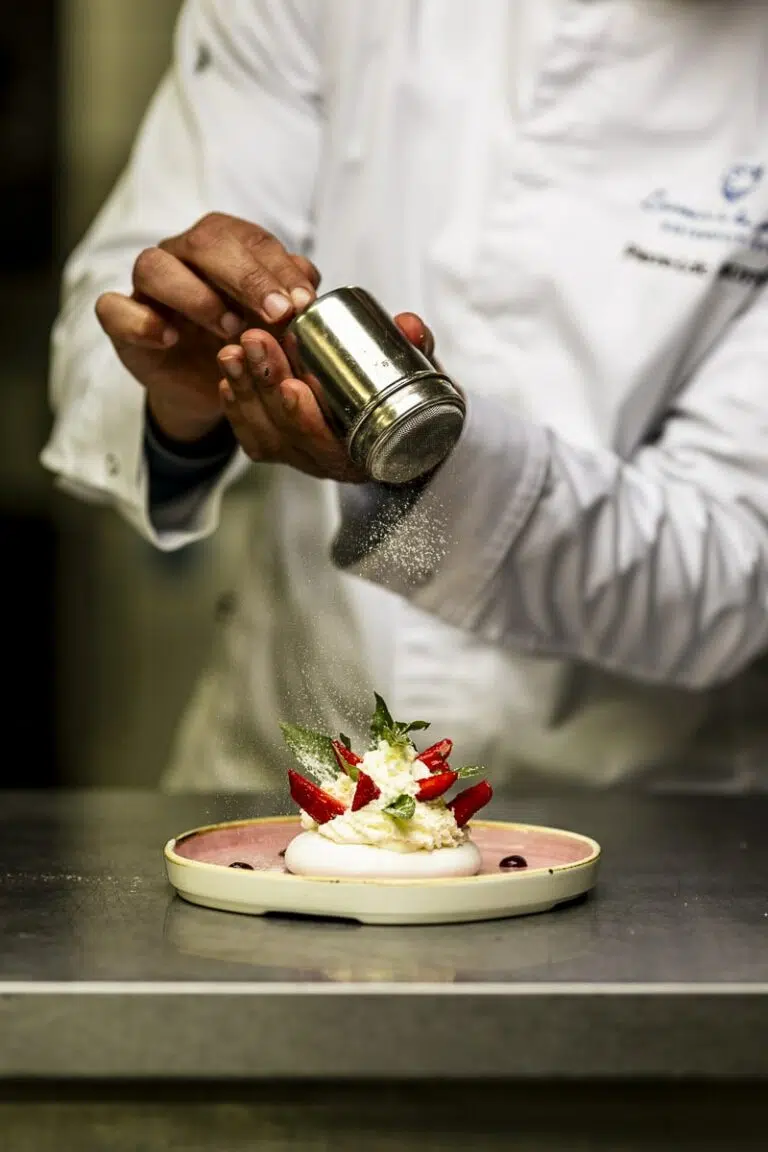 Un chef en blouse blanche saupoudre du sucre glace sur une pavlova garnie de crème chantilly, fraises et feuilles de menthe, dans une cuisine professionnelle.