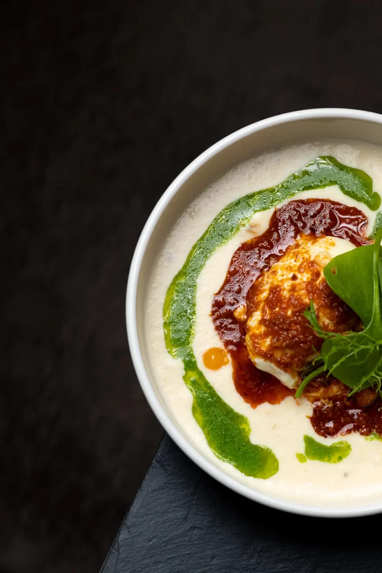 Pièce rôtie dorée, sauce brun-rouge brillante, émulsion blanche et cordon d'herbes vertes dans une assiette creuse blanche, fond ardoise.